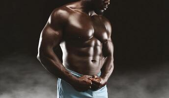 A muscular man posing in studio, displaying defined abs and biceps in shorts against dark background for dramatic effect. photo