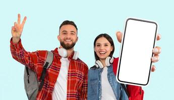 A young man and woman stand in front of a light blue background. The man is wearing a red plaid shirt and a backpack and has headphones around his neck. photo