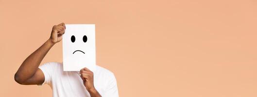 A man is holding a white sign with a sad face drawn on it in front of his face. The sign is held up with his left hand, while his right hand is resting on the sign, copy space photo