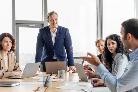 A group of business professionals are gathered around a table, discussing ideas. One man is standing, looking at the group while another man gestures, engaged in the conversation photo