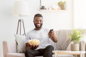 Black man is sitting on a couch in a living room. He is holding a bowl of popcorn and a remote control in his hands. He is smiling and looking at the television photo