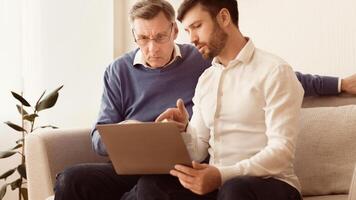 Mature man teaching old dad to use laptop, computer programs and browse internet sitting on sofa indoor. Panorama photo
