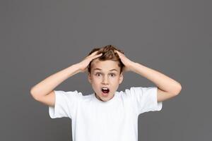 A shocked teen boy is captured with wide eyes and hands clutching his head against an isolated grey backdrop, suggesting alarm photo