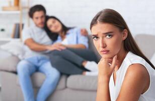 Jealousy. Unhappy Girl Looking At Camera While Her Dating Friends Hugging Sitting On Sofa Indoor. Selective Focus photo