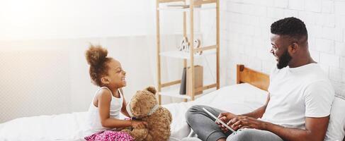 A little black girl is sitting on a bed, hugging a teddy bear and smiling at her dad. Her dad is sitting next to her, using a tablet and smiling back at her. photo