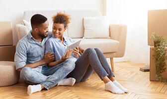 Relocation Concept. Joyful Black Spouses Using Tablet Computer Sitting On Floor Among Moving Boxes In New House. Copy Space photo
