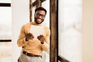 Happy Black Man Holding Tablet , Standing In Front Of The Big Window At Office, Looking At Camera. Empty space photo