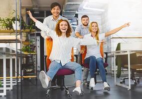 Time for break. Happy colleagues having fun at workplace, making office chair race in coworking space photo