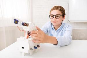 A young boy sits at a desk in a classroom, focusing intently as he assembles a robotic arm kit. The focus on his face suggests his interest in STEM education and the excitement of creating photo