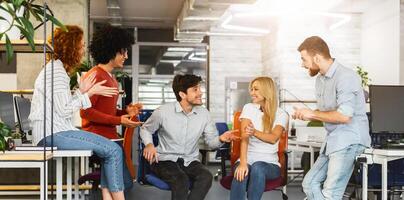 A group of young professionals is gathered in a brightly lit office space, engaging in an animated discussion. Some members are seated while others stand, indicating collaborative work environment photo