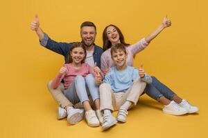 A smiling family consisting of a mother, father, daughter, and son are sitting close to each other against a vibrant yellow backdrop, all giving a thumbs-up sign in a show of happiness and unity. photo