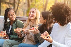 Cheerful multiracial teens joking and laughing while eating pizza, sitting on bench in park photo