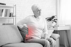 Senior woman suffering from backache, sitting on couch and touching loin at home, empty space photo