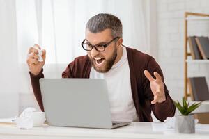 A bearded man with glasses is sitting in a bright office, visibly frustrated and shouting at his laptop. Papers and a plant are on the desk, suggesting a working environment. photo