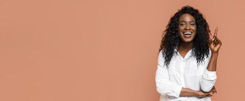 Smiling Black woman with long, curly hair. She is wearing a white shirt and pointing upwards with her right index finger. The background is a simple peach color. photo