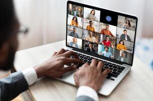 Black man sits at a table in front of an open laptop, actively participating in a conference. The laptop screen displays a grid of various participants, each in their own frame photo