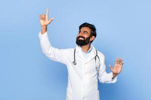 Indian man doctor with a beard is smiling and pointing upward with his right hand. His left hand is outstretched with his palm facing forward. He is wearing a white lab coat with a stethoscope. photo