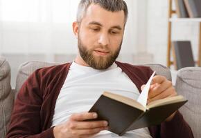 A man is seated on a couch, engrossed in reading a book. He is focused and turning the pages, with a concentrated expression on his face. The room is well-lit, creating a cozy reading environment. photo