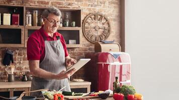 A senior man in a red shirt and apron stands in his kitchen, holding a tablet and smiling. He is following a recipe while surrounded by fresh vegetables, a cutting board, and a red refrigerator photo
