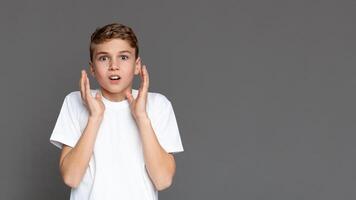 A teenager with a shocked expression holds his cheeks isolated against a gray background, showing a range of youngster's emotions, copy space photo