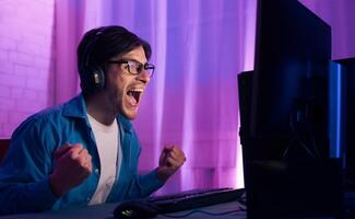 A young man wearing headphones and glasses is seated in front of a computer screen playing games. He is excited and has his arms raised in the air as if in victory. photo