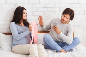 Gadget addiction concept. Indifferent husband ignoring his pregnant wife, using smartphone in bed while woman showing him clothes for future baby photo