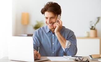 A man is smiling and talking on a phone while working on a laptop. He is seated at a desk and has paperwork in front of him. photo