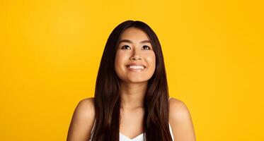 A young Asian woman with long dark hair is smiling as she looks up towards the sky. She is wearing a white tank top and standing in front of a solid yellow background. photo