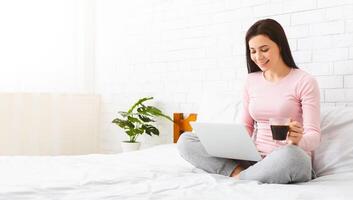 A woman is seated on a bed, working on a laptop while holding a cup of coffee. She appears focused and engaged in her work, with a cozy and relaxed atmosphere surrounding her, empty space photo