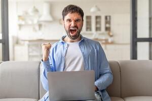 A man with a beard sits on a beige couch in a living room and excitedly celebrates with a raised fist, looking directly at the camera while using a laptop. photo