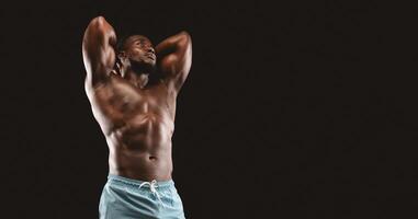 A muscular man with a defined physique poses with his arms behind his head, looking up, against a black background. photo
