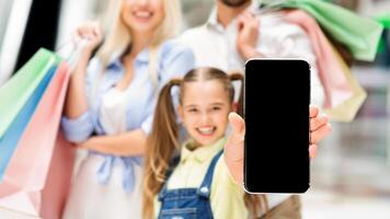 A smiling young girl holds a smartphone with a blank screen towards the camera. Her parents are blurred in the background, holding shopping bags. photo
