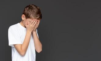 The young boy in a white tee covers his eyes with hands, expressing fear, shame, or a desire not to see, isolated on a grey background photo