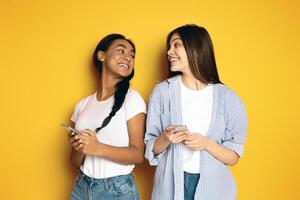 Two multiethnic girls one with a braid and the other with long hair, stand side by side against a vibrant yellow backdrop, sharing a joyful moment. Each holds a smartphone photo