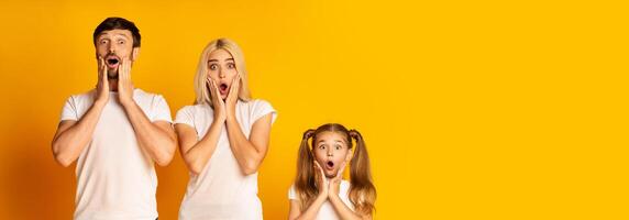 A family is displaying expressions of shock and excitement against a vibrant yellow backdrop. The adults and child appear astonished, with hands on their faces and wide eyes, copy space photo