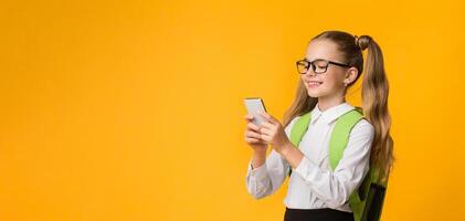 Cheerful School Girl Using Smartphone On Yellow Studio Background. Free Space For Text photo