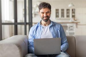 A man sits on a couch in a modern living room and uses a laptop. He is wearing a blue shirt, and he is smiling as he looks down at the screen. There are glass doors in the background. photo