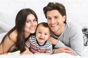 This image depicts a happy family of three - a mother, father, and their young baby - lying on a white bed together. The parents are smiling and looking at the camera photo