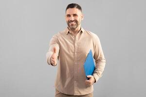 A well-dressed man smiles warmly as he extends his hand for a handshake. He holds a blue folder in his other hand, which may contain important documents. The background is plain and grey photo