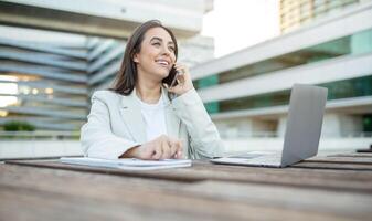 A young businesswoman sits outside, working on her laptop and taking a phone call. She is smiling and appears to be enjoying her work. The background features an office building with large windows. photo