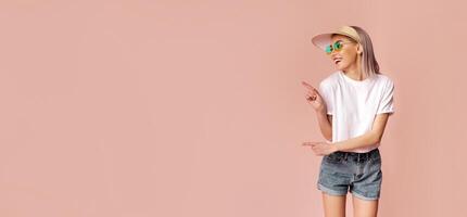 A woman in sunglasses and a hat is standing in front of a pink background. She is smiling and pointing to the left with her right hand, copy space photo