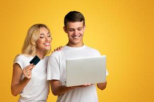 Young couple shopping online with laptop and credit card over yellow studio background, copy space photo
