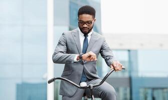 A Black businessman in a suit is riding a bicycle and checking his watch in an urban setting. The scene is modern and bustling, highlighting time management and a busy lifestyle. photo