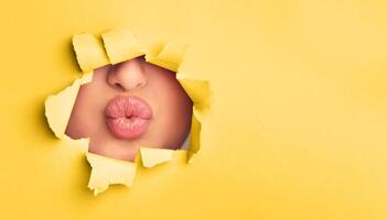 A close-up view of a woman lips subtly peeking out of a hole in a bright yellow paper. The lips are painted in a bold red color, adding a striking contrast to the paper. photo