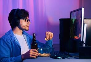 This image shows a young man playing games while wearing headphones. He is sitting in front of a computer with a controller in his hand. He is enjoying snacks and a beer while he plays. photo