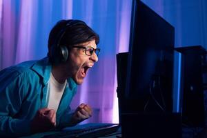 A young man wearing headphones and glasses sits at his computer and screams with excitement while playing a game. The room is dimly lit, and the monitor is brightly lit photo