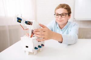 A young boy wearing glasses is seen at a desk, focused on assembling a white robotic arm model. He carefully aligns the components, demonstrating his interest in robotics and STEM learning. photo