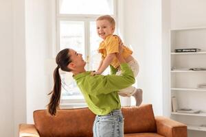 A woman cheerfully holds a child up in the air, both smiling. The child arms are outstretched, enjoying the playful moment. Mother face shows joy and love for her son photo