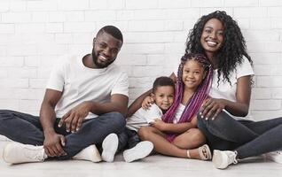 Joyful afro family mother, father and two little children sitting on floor and cuddling over white brick wall background. photo