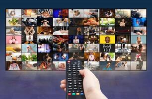 A hand is holding a black remote control in front of a television screen that is displaying a variety of channels. The screen is filled with a grid of small squares photo